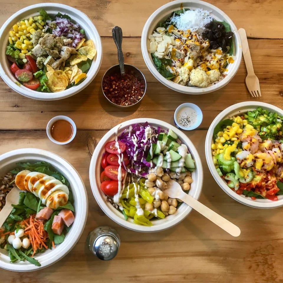 Five vibrant Leaf salads in white bowls, with various toppings and dressings, on a wooden table.